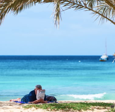Homem relaxa numa praia de Cabo Verde, lendo um jornal sob a sombra de palmeiras, com o mar azul e um barco ao fundo.